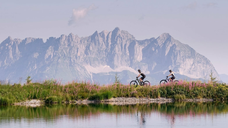Zwei Mountainbiker mit E-Mountainbikes an einem See vor einer beeindruckenden Bergkulisse.