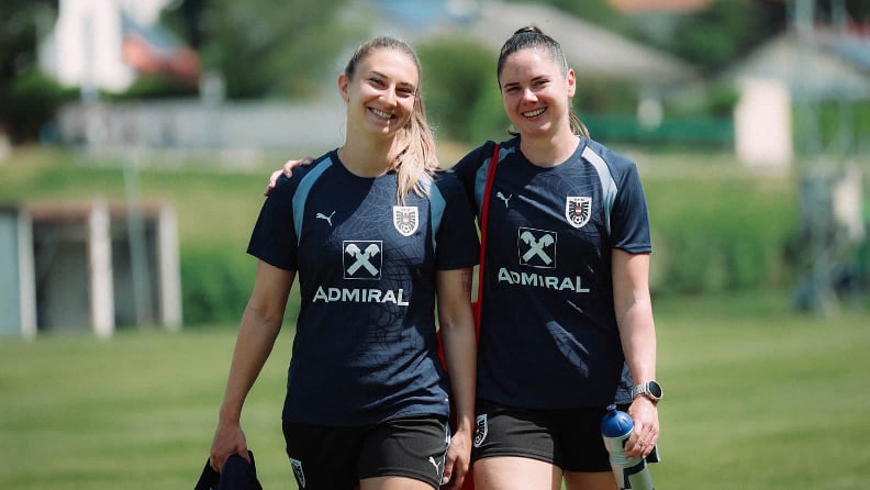 Zwei Sportlerinnen in Trainingskleidung gehen nebeneinander über einen Rasenplatz.