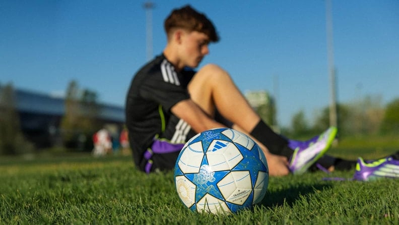 Fußball liegt im Vordergrund auf Rasen, während ein Spieler im Hintergrund sitzend seine Schuhe anzieht. Der Fokus liegt auf dem Ball in Bodennähe.
