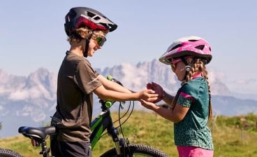 Zwei Kinder mit Fahrradhelmen stehen neben einem Fahrrad und halten sich lachend die Hände, im Hintergrund sind Berge zu sehen.