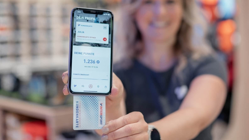 Eine Frau hält ein Smartphone mit geöffneter INTERSPORT-App, die Kundenkarte und Punkte anzeigt, sowie eine physische Kundenkarte.