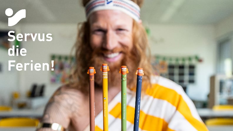 Vier bunte Stifte mit Figurenköpfen vor einer lächelnden Person, dazu der Schriftzug Servus die Ferien!