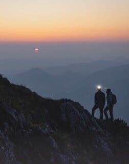 Zwei Personen mit Stirnlampen stehen auf einem Berggrat bei Sonnenuntergang.