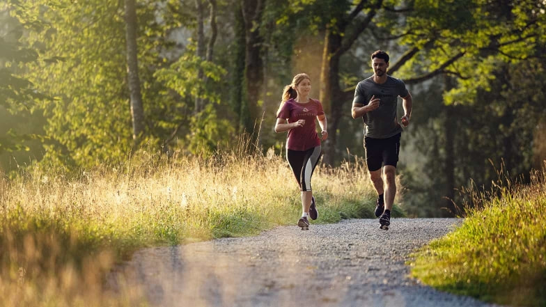 Eine Läuferin und ein Läufer beim gemütlichen Abendlauf. Das Licht der untergehenden Sonne lässt die Wiese neben dem Weg leuchten.