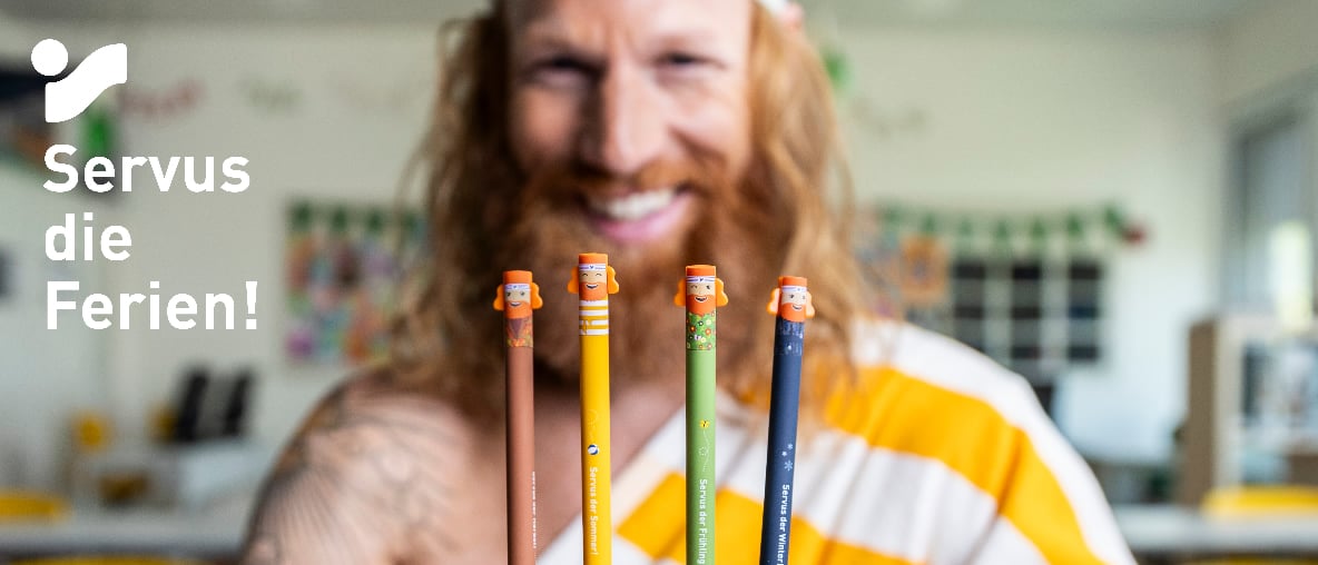 Vier bunte Stifte mit Figurenköpfen vor einer lächelnden Person, dazu der Schriftzug Servus die Ferien!