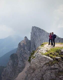 Zwei Personen mit Rucksäcken stehen auf einem Felsen mit Blick auf steile Bergspitzen.