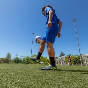 Fußballspieler steht auf einem Kunstrasenplatz und kontrolliert einen Ball mit dem Fuß. Im Hintergrund sind ein Tor und weitere Spieler zu sehen.
