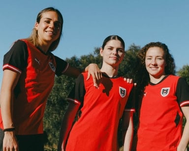 Drei Frauen tragen rot-schwarze Fußballtrikots und stehen eng beieinander im Freien. Sie lächeln in die Kamera vor blauem Himmel.