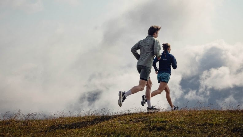 Zwei Läufer joggen über einen Bergrücken, umgeben von Wiesen und tief hängenden Wolken.