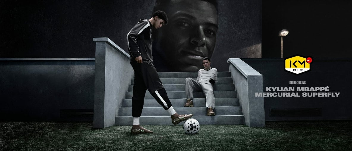 Zwei Männer mit Fußball vor einem riesigen Mbappé-Wandbild.