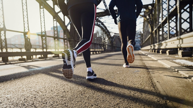 Zwei Personen joggen bei sonnigem Wetter über eine Stahlbrücke.