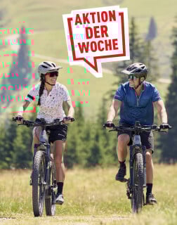 Lächelnde Radfahrer auf Mountainbikes in einer grünen Landschaft mit Bäumen im Hintergrund, daneben eine rote Sprechblase mit der Aufschrift „Aktion der Woche“.