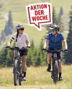 Lächelnde Radfahrer auf Mountainbikes in einer grünen Landschaft mit Bäumen im Hintergrund, daneben eine rote Sprechblase mit der Aufschrift „Aktion der Woche“.