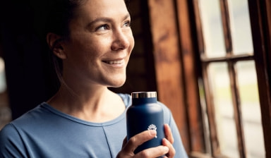 Eine lächelnde Frau mit blauer Flasche von Hydroflask schaut aus dem Fenster.