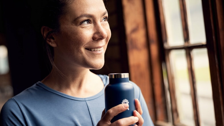 Eine lächelnde Frau mit blauer Flasche von Hydroflask schaut aus dem Fenster.