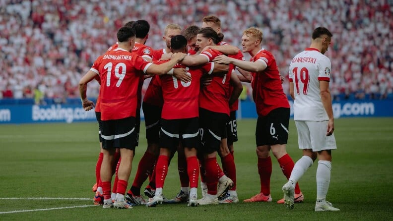 Mehrere Fußballspieler in roten Trikots stehen eng zusammen und umarmen sich auf dem Spielfeld. Ein Spieler in weißem Trikot steht daneben und blickt zur Seite.