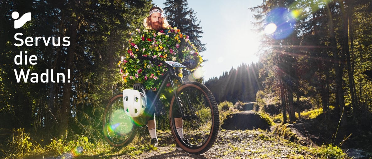 Radfahrer mit Blumenkostüm auf Mountainbike im Wald bei Sonnenlicht, Text „Servus die Wadln!“
