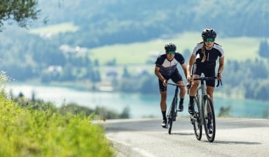 Eine Frau und ein Mann fahren mit dem Rennrad einen Berg hinauf. Hinter ihnen sieht man in der Ferne im Tal einen See.