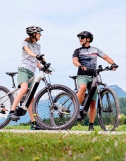 Frau und Mann stehen mit E-Mountainbikes auf einer Wiese und blicken sich an, Berglandschaft im Hintergrund.