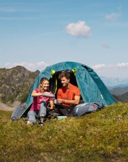 Zwei Personen sitzen in Schlafsäcken vor einem aufgebauten Zelt in einer Berglandschaft.