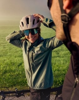 Frau mit Fahrradhelm und Sonnenbrille richtet ihren Helm, während sie neben einem Fahrrad auf einer Wiese steht.