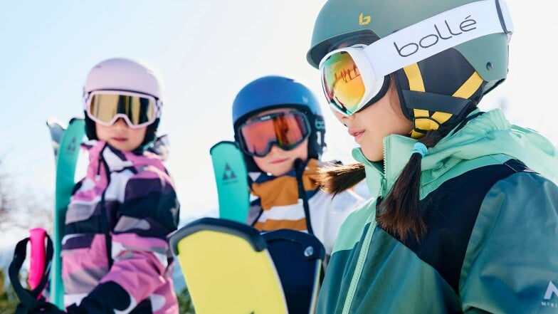 Drei Kinder mit Skihelmen und Brillen stehen im Schnee.