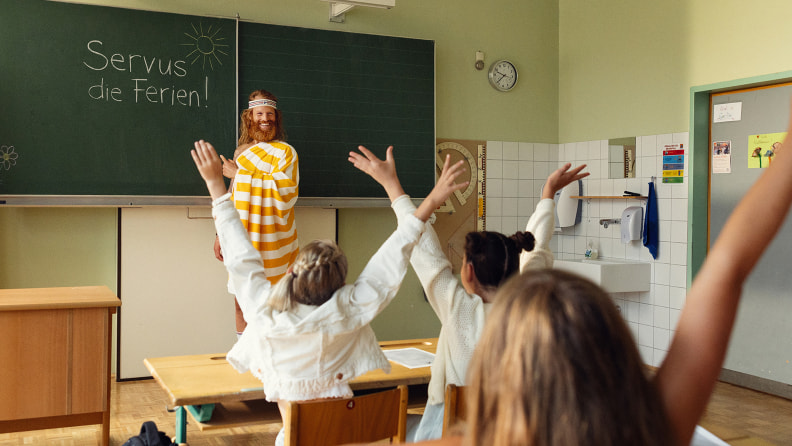 Der Sommer (Mann mit Bart und luftigem, gelbgestreiftem Umhang) steht in einer Schulklasse und zeigt auf die Tafel, auf der "Servus, die Ferien!" steht. Die Kinder jubeln.