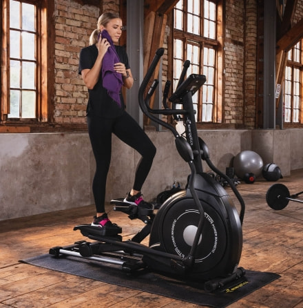 Eine Frau trainiert auf einem Crosstrainer von Energetics in einem lichtdurchfluteten Raum mit Hantelbänken und Gymnastikbällen.