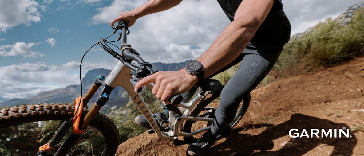 Garmin-Smartwatch am Handgelenk einer Person auf einem Mountainbike auf einem unbefestigten Weg vor Bergen