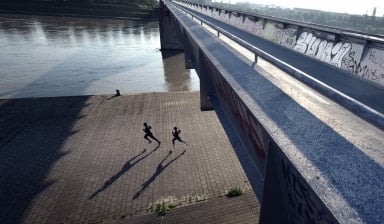 Zwei Läufer laufen mit langen Schatten unter einer Brücke am Fluss. Es ist früh am Morgen.
