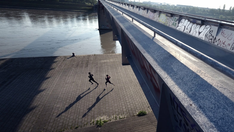 Zwei Läufer laufen mit langen Schatten unter einer Brücke am Fluss. Es ist früh am Morgen.