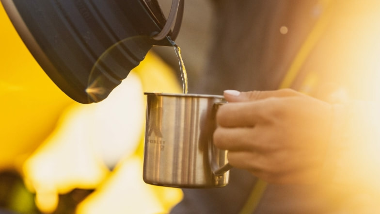 Eine Hand gießt heißes Wasser in eine metallene Camping-Tasse.