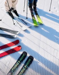 Mehrere Paar Ski liegen nebeneinander auf frisch präpariertem Schnee, zwei Personen stehen mit Skiern bereit.