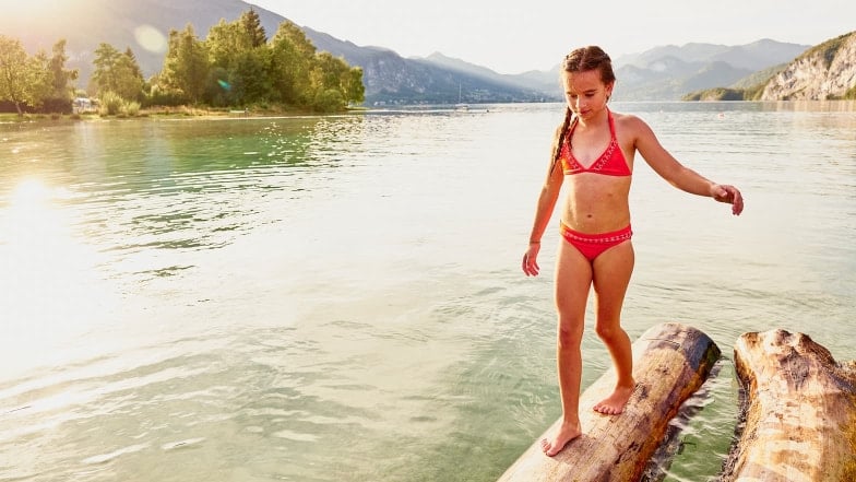 Ein Mädchen balanciert am Ufer eines Sees über einen im Wasser liegenden Baumstamm.