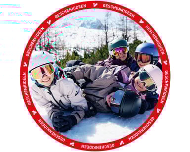 Vier lachende Kinder mit Skiausrüstung liegen verspielt im Schnee.