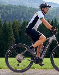 Mann mit Helm fährt ein E-Mountainbike auf einem Weg vor bewaldeten Bergen.