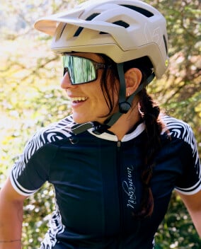 Radfahrerin mit weißem Helm und Sonnenbrille, lächelnd beim Training im Grünen bei warmem Tageslicht