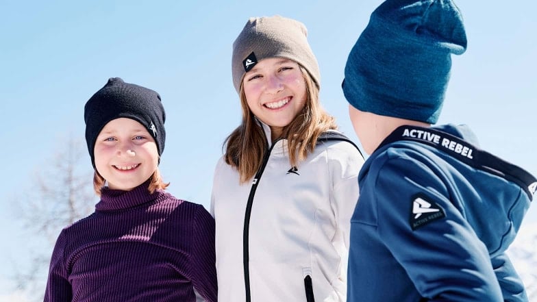 Drei glückliche Kinder mit Hauben stehen draußen bei Sonnenschein.