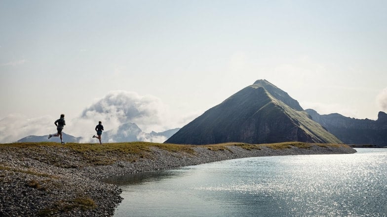 Zwei Personen laufen an einem See in einer Berglandschaft entlang.