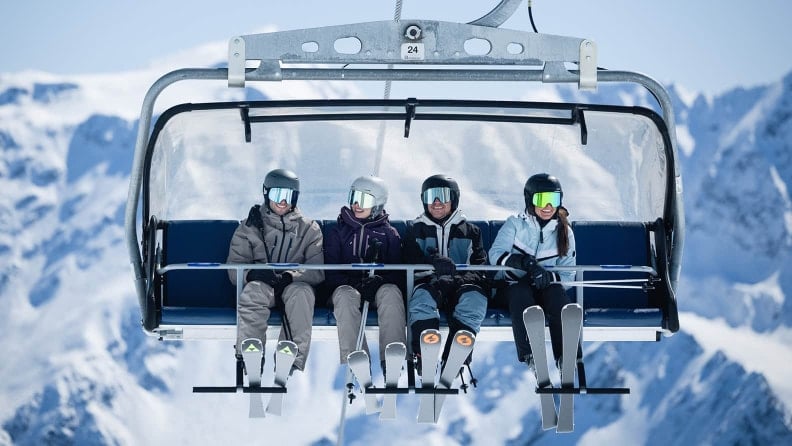 Vier Personen mit Skiausrüstung sitzen nebeneinander in einem Sessellift.