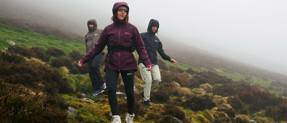 Drei Menschen wandern in Regenjacken von Jack Wolfskin durch eine neblige Hügellandschaft.