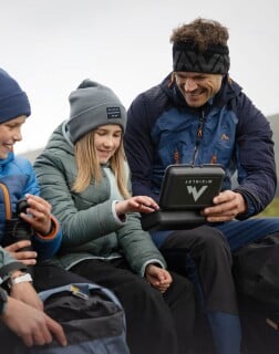 Ein Mann und zwei Kinder in Outdoor-Kleidung sitzen zusammen und machen eine Jausenpause.