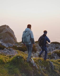 Zwei Wandernde mit Rucksäcken gehen über felsiges Gelände im Abendlicht.