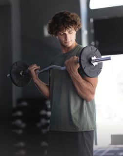 Mann führt im Stehen Bizepscurls mit einer Langhantel aus. Die Übung dient dem Aufbau der Armmuskulatur.
