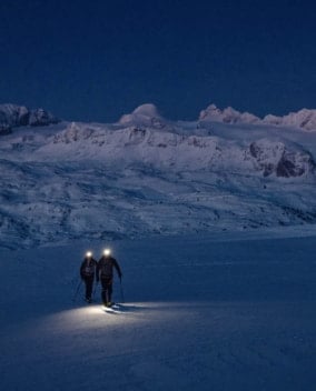 Zwei Skitourengeher mit Stirnlampen steigen in der Dämmerung durch verschneite Berglandschaft.