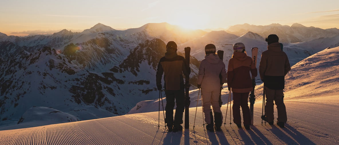 Vier Skifahrerinnen und Skifahrer stehen bei Sonnenuntergang auf einer präparierten Skipiste vor verschneiten Bergen