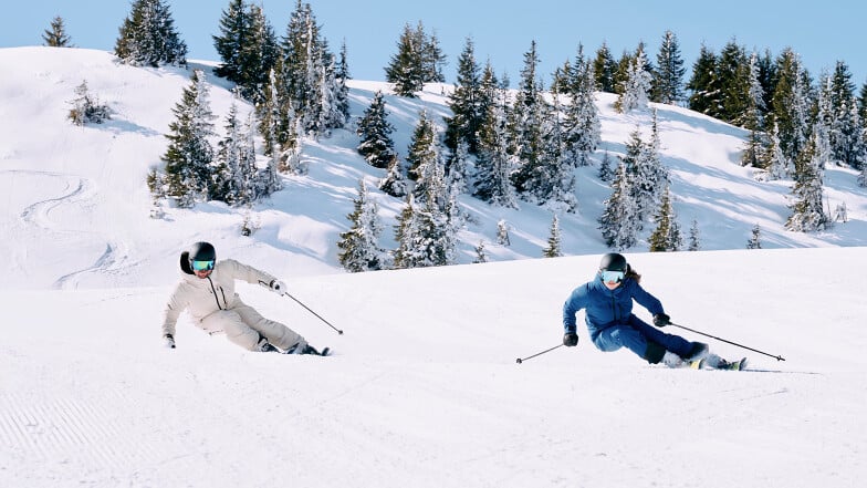 Zwei Skifahrer auf einer sonnigen, verschneiten Piste in den Bergen.