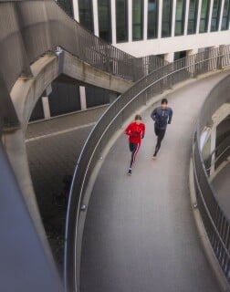 Zwei Personen joggen nebeneinander auf einer geschwungenen Rampe zwischen Gebäuden.