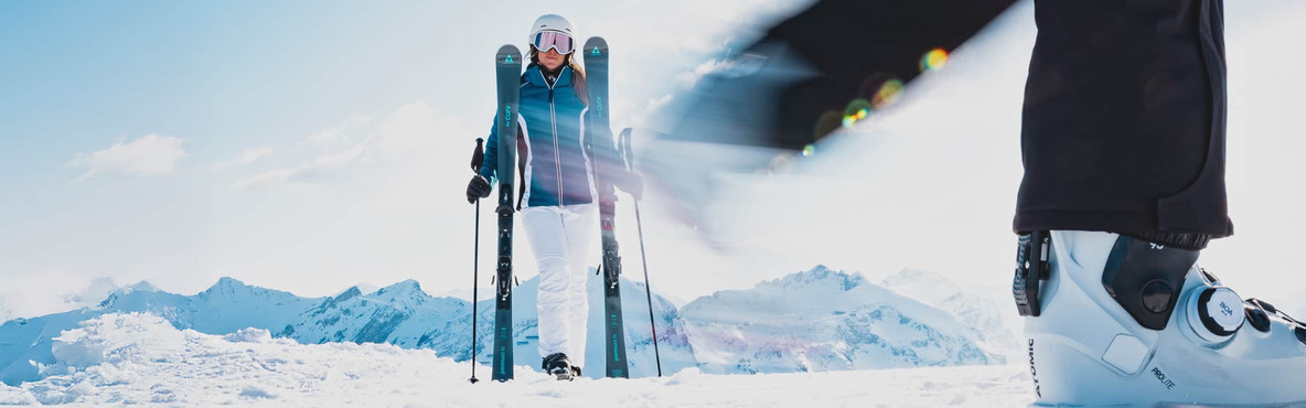 Eine Skifahrerin trägt Ski und Stöcke durch eine verschneite Berglandschaft.