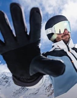Eine Person mit Skihelm und Handschuh streckt die Hand zur Kamera aus, im Hintergrund verschneite Berge.